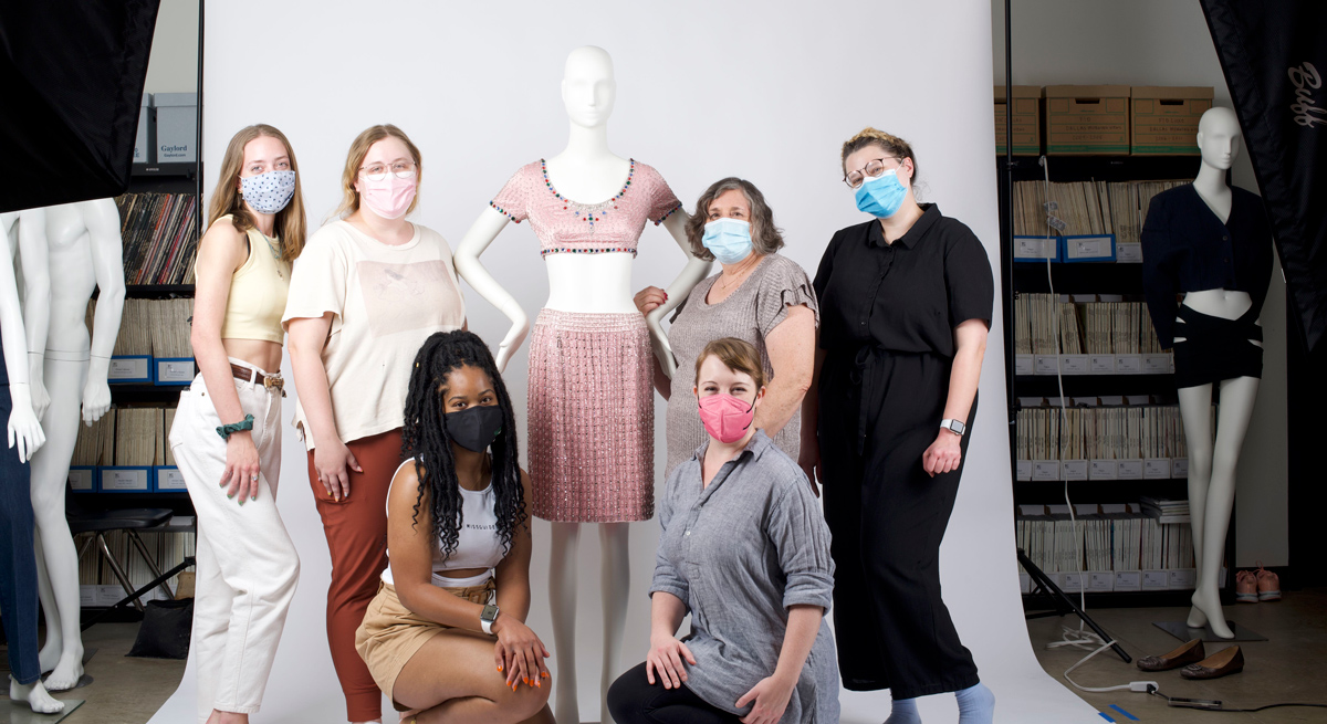 Six people stand in front of a white photo backdrop around a mannequin showing a pink ensemble; in the background are bookshelves and more mannequins, in the foreground lighting equipment.
